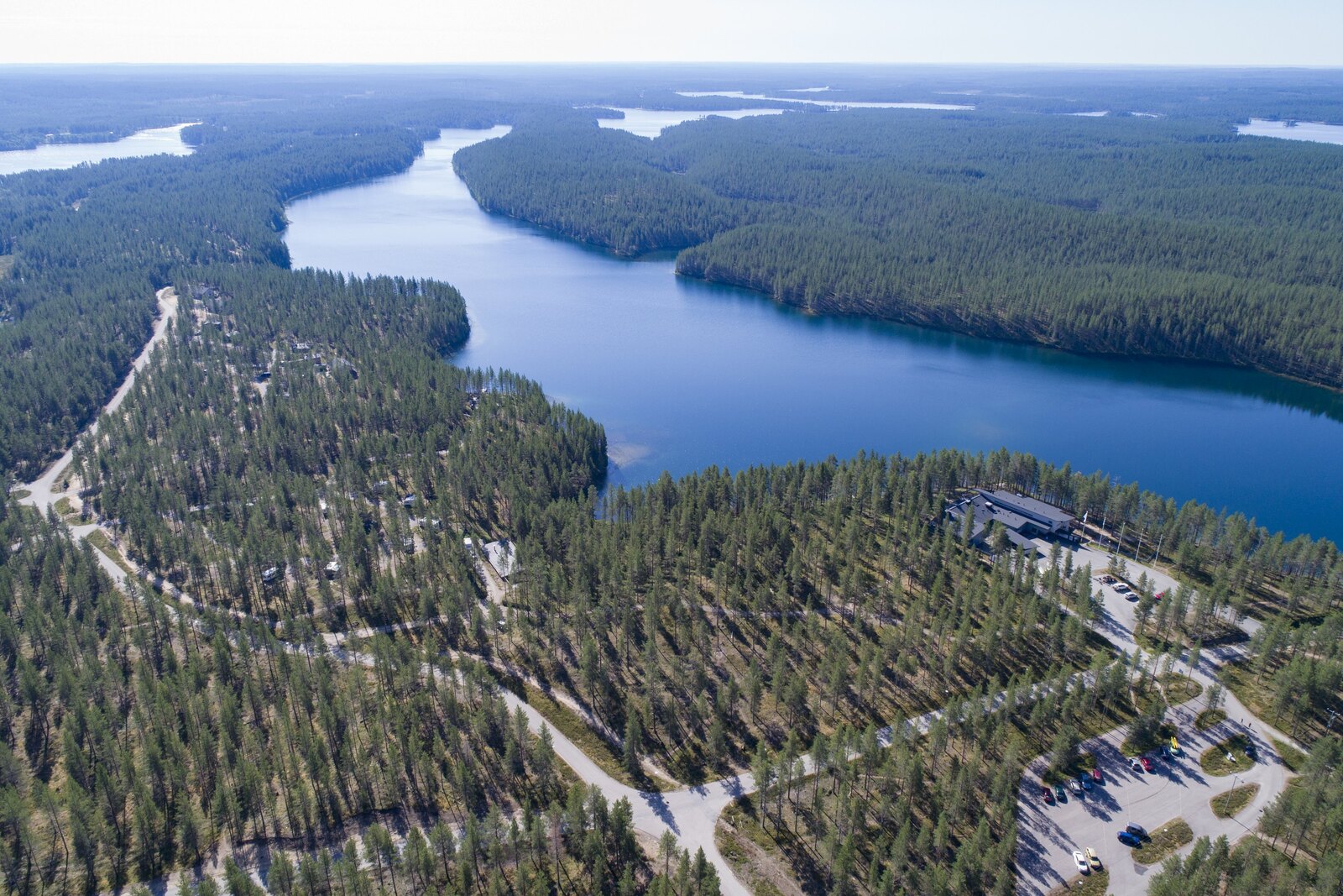Horisonttiin jatkuva metsä reunustaa järveä. Etualalla tiet risteilevät mökkialueella. Ilmakuva.