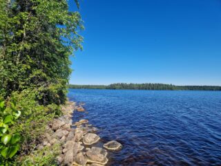 Rannan kiviä ja lehtipuustoa, kaukana näkyy vastarantaa.