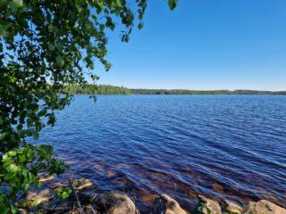 Rannan kivikkoa, vastaranta näkyy kaukana, lehtipuut työntyvät kuvaan.