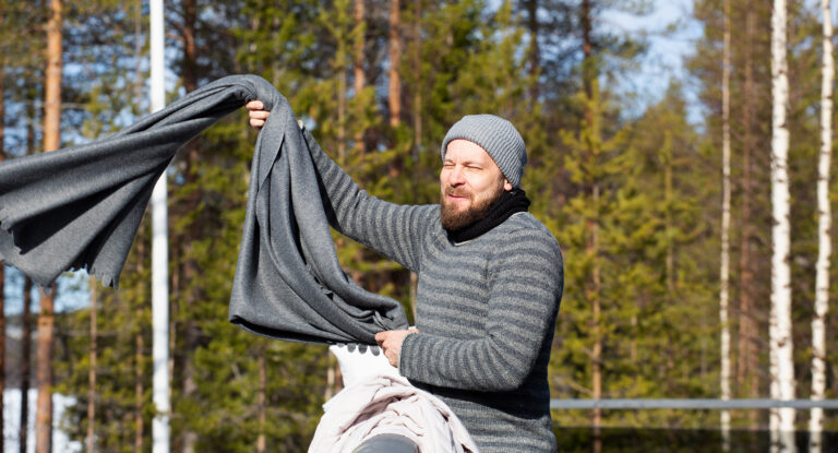 Mies harmaassa pipossa ja toppatakissa tuulettaa vuodevaatteita kevättalvisen mökin portailla. On juuri heittämässä harmaata vilttiä kaiteen yli.