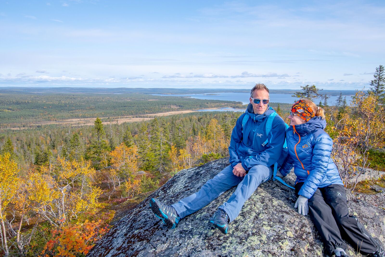 Kaksi henkilöä istuvat ison kiven päällä tunturin laella. Maisemassa näkyy ruskasävyjä.