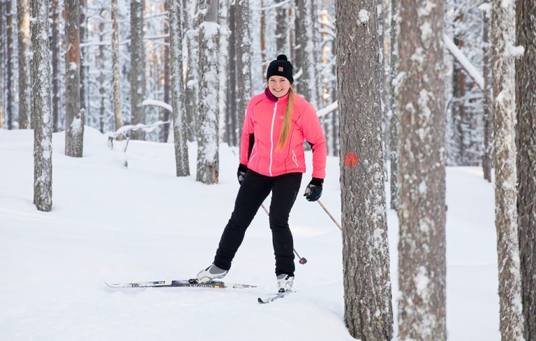 Kirkkaan persikanväriseen takkiin pukeutunut luisteluhiihtäjä laskee loivaa rnnettä mäntymetsässä ja katsoo hymyillen kameraan.