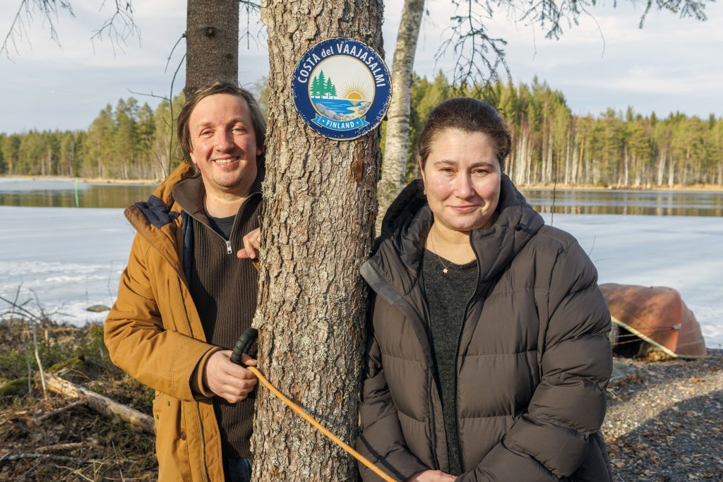 Kaksi henkilöä nojaa puuhun kahdelta puolen. Taustalla näkyy keväinen, osittain jään peittämä järvimaisema.