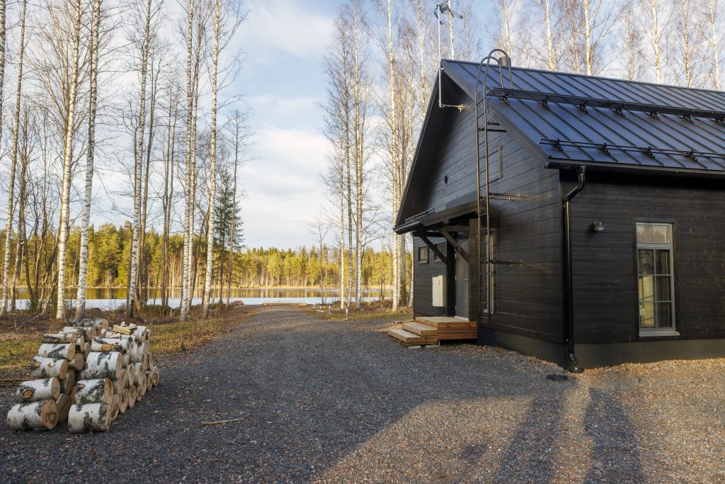 Pihatie vie tumman asuintalon ohitse rantaan. talven jäljiltä puut ovat paljaina, järvi on vapautunut jäästä.