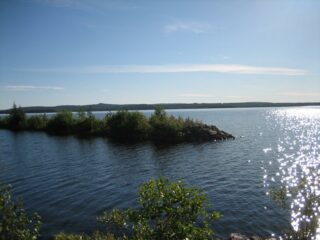 Kesämaisemassa lähellä järven rantaa on aallonmurtaja. Vesi kimmeltää auringonvalossa.