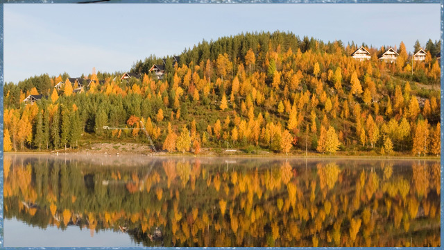 Järven rannasta kohoavassa rinteessä on mökkejä. Ruskansävyinen metsä heijastuu järven pintaan.