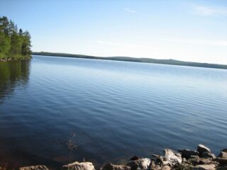 Kesämaisemassa pilvetön taivas ja vasemman laidan metsä heijastuvat järveen. Etualalla on kiviä.