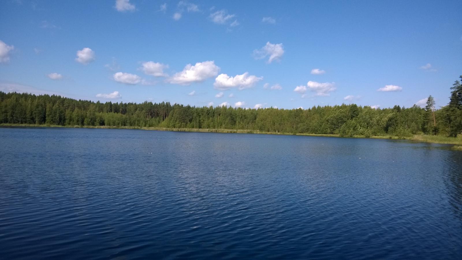 Kesäisessä järvimaisemassa veden pinta väreilee hieman. Taivaalla on muutamia poutapilviä.