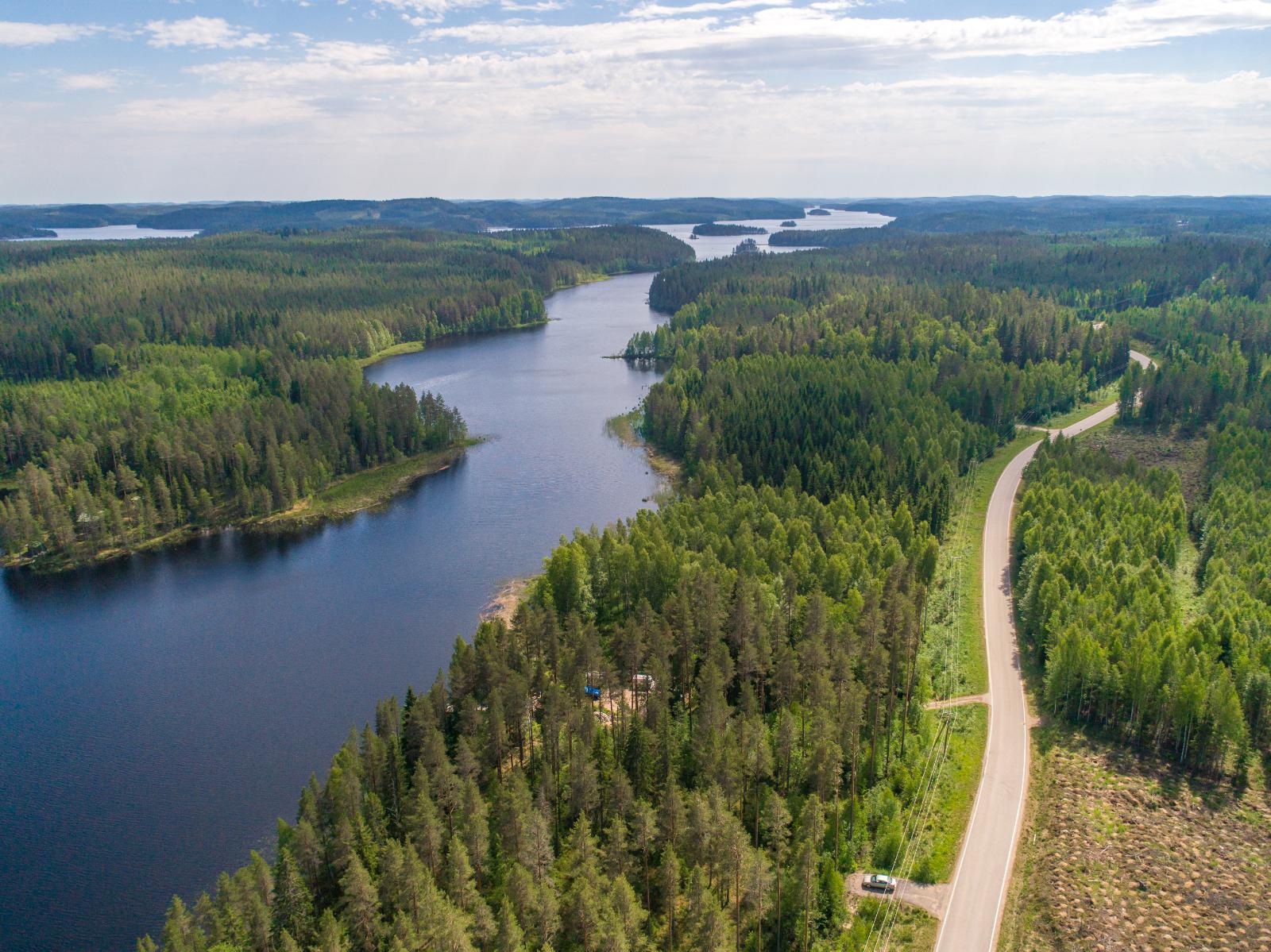 Metsät reunustavat horisonttiin jatkuvaa pitkulaista järveä. Vasemmalla kulkee tie. Ilmakuva.