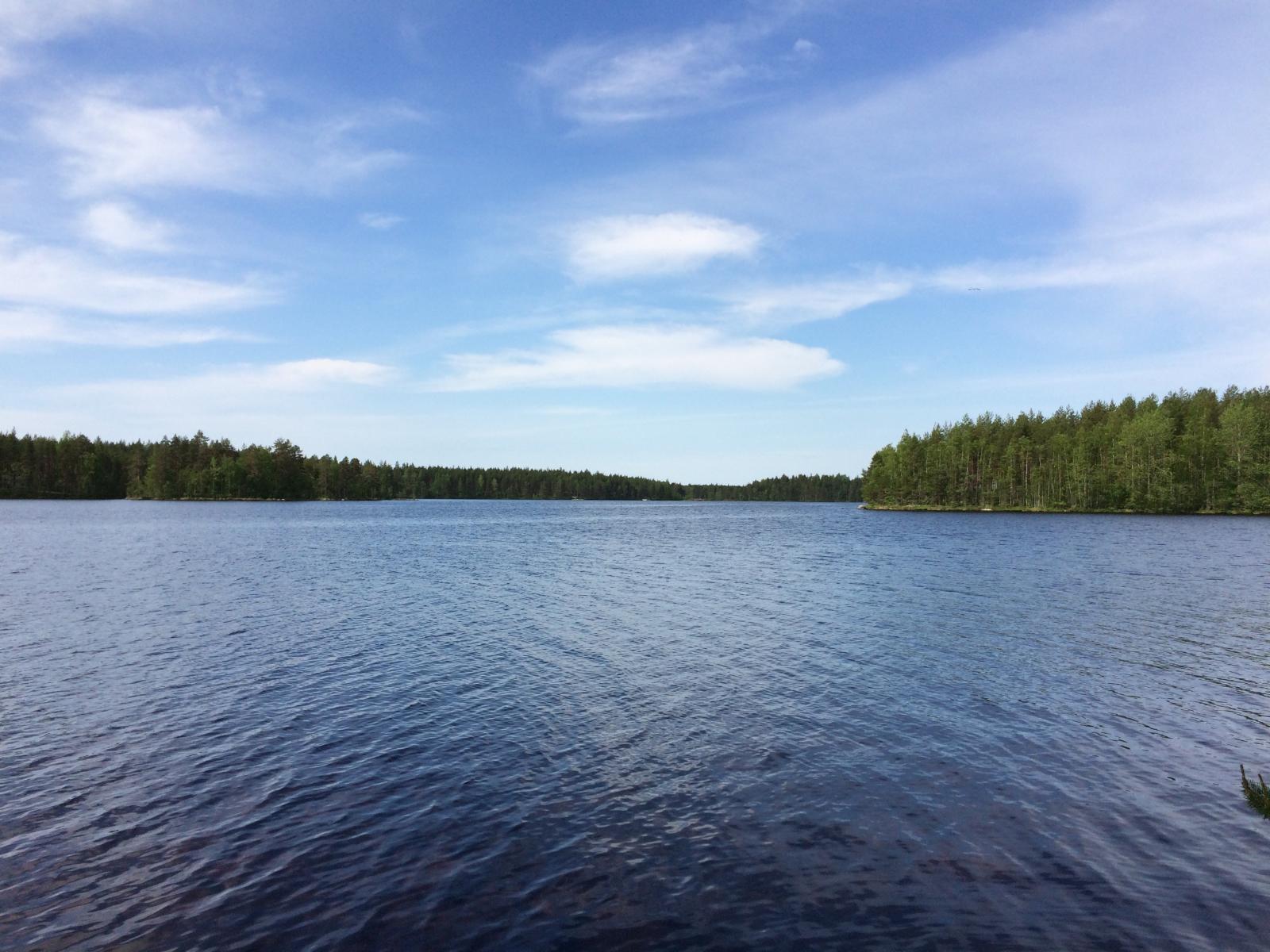 Kesämaisemassa puolipilvinen taivas heijastuu hieman väreilevään järveen. Vastarannalla metsää.