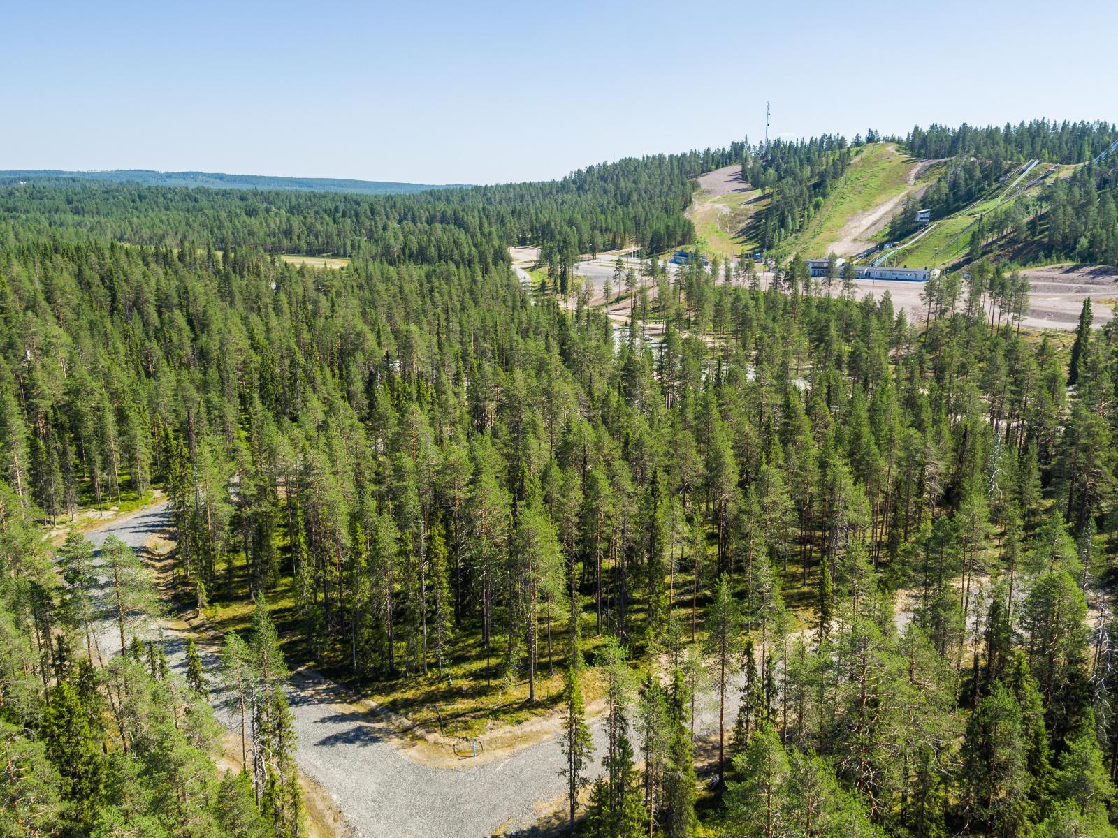 Kesäisessä ilmakuvassa tiet risteävät havumetsässä. Taustalla laskettelurinteet ja pysäköintialue.