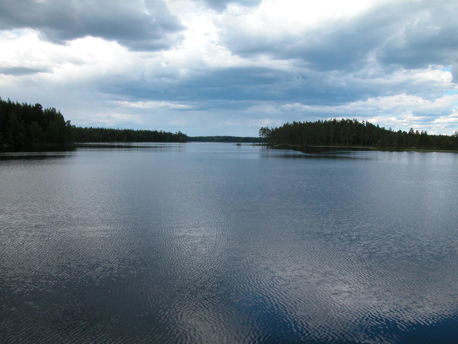 Poutapilvinen taivas heijastuu järven hieman väreilevään pintaan. Järven astarannalla on metsää.