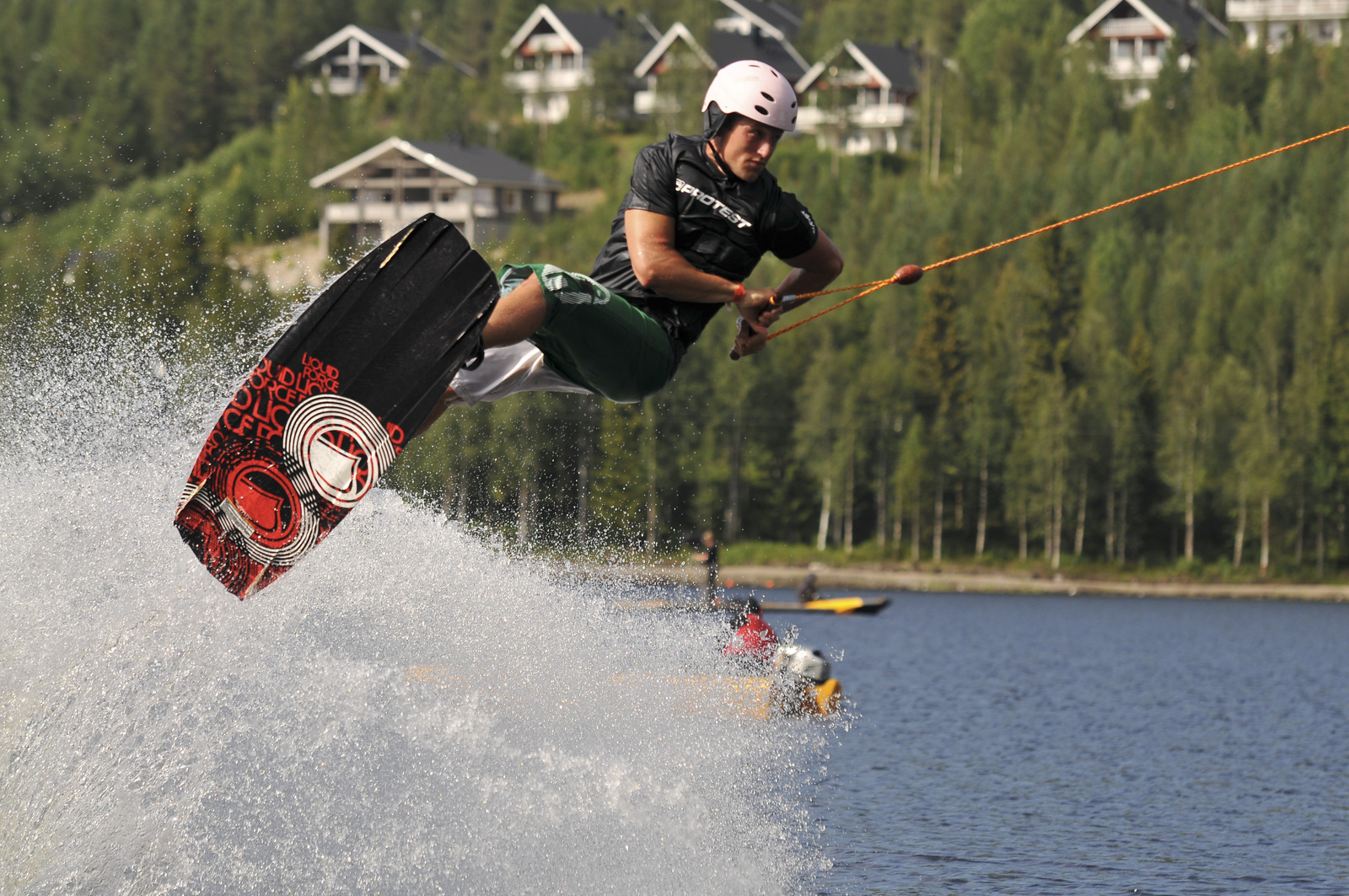 Wakeboard-harrastajan ilmatempussa vesi roiskuu järvellä. Taka-alalla metsäisen rinteen mökkialue.