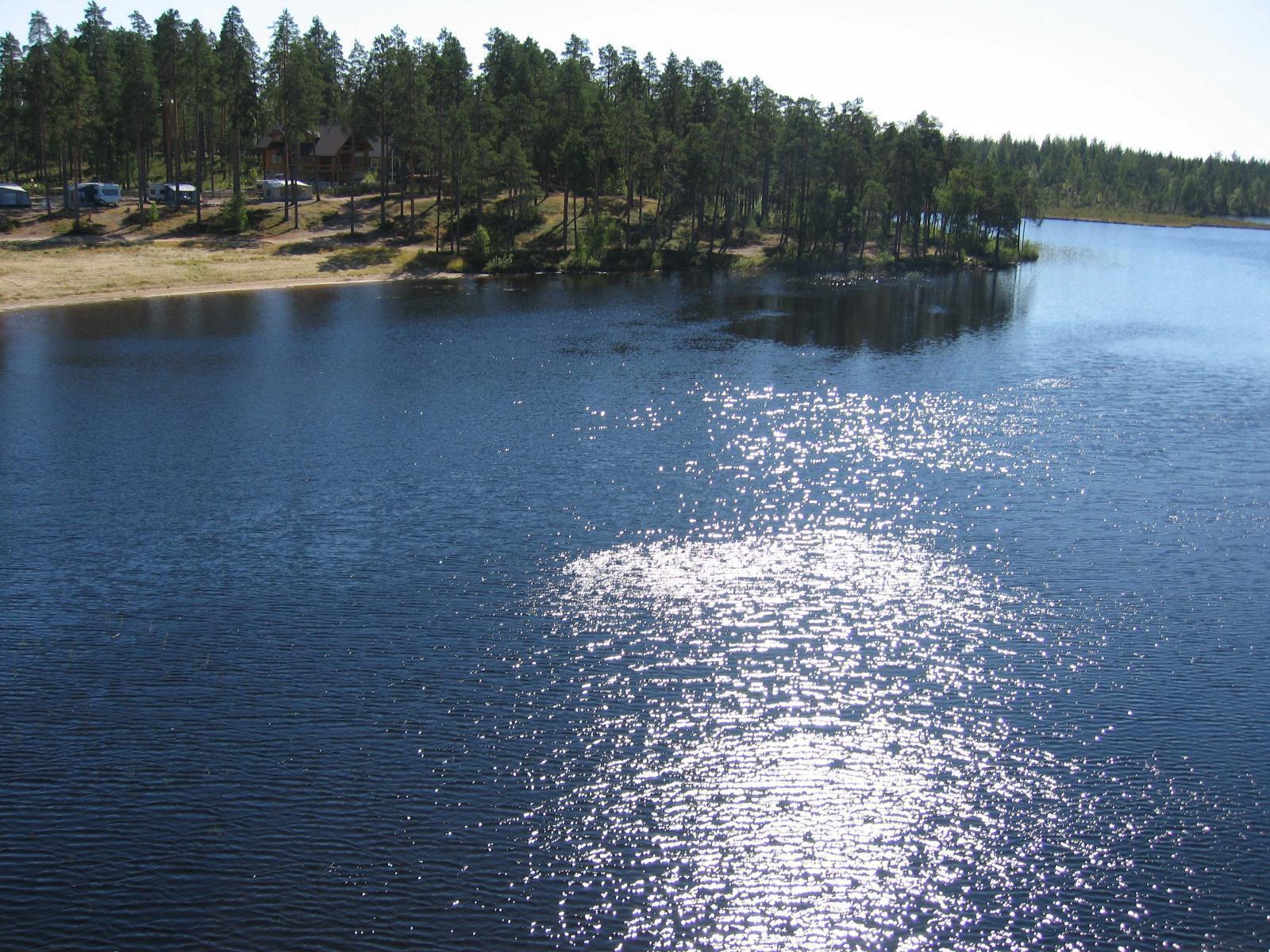 Järven vastarannalla on hiekkaranta ja leirintäalue. Vedenpinnasta heijastuu auringonvalo. Ilmakuva.