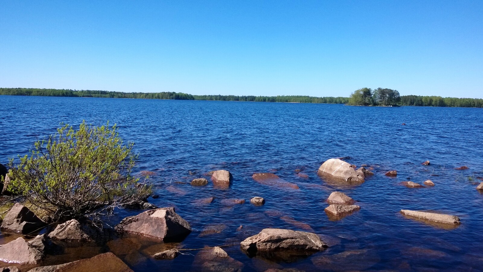 Kesäisessä järvimaisemassa veden pinta laineilee hieman ja rantavedestä pilkistää kiviä.