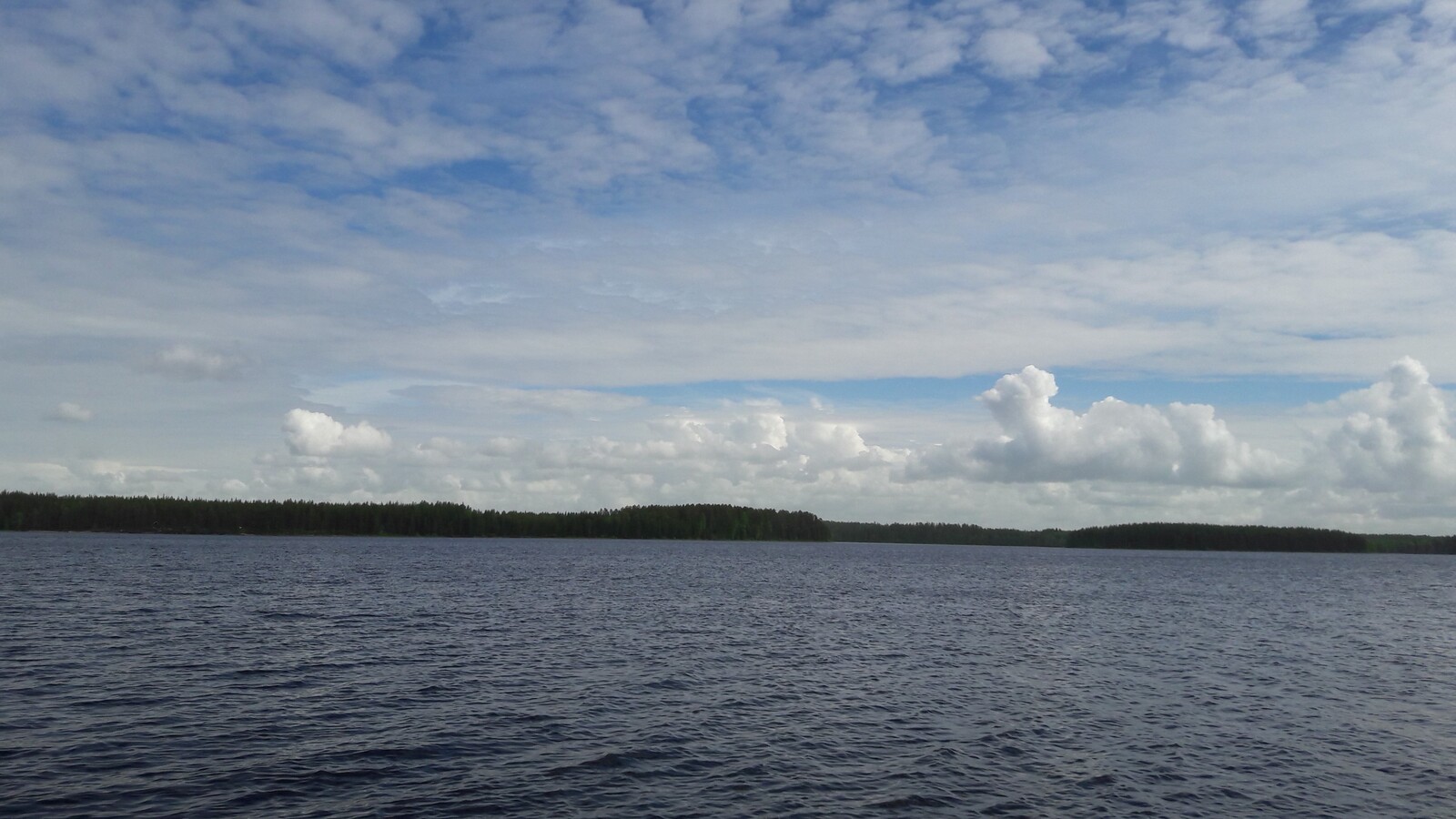 Laajassa järvimaisemassa vesi laineilee hieman. Taivalla on erilaisia pilviä eri kerroksissa.