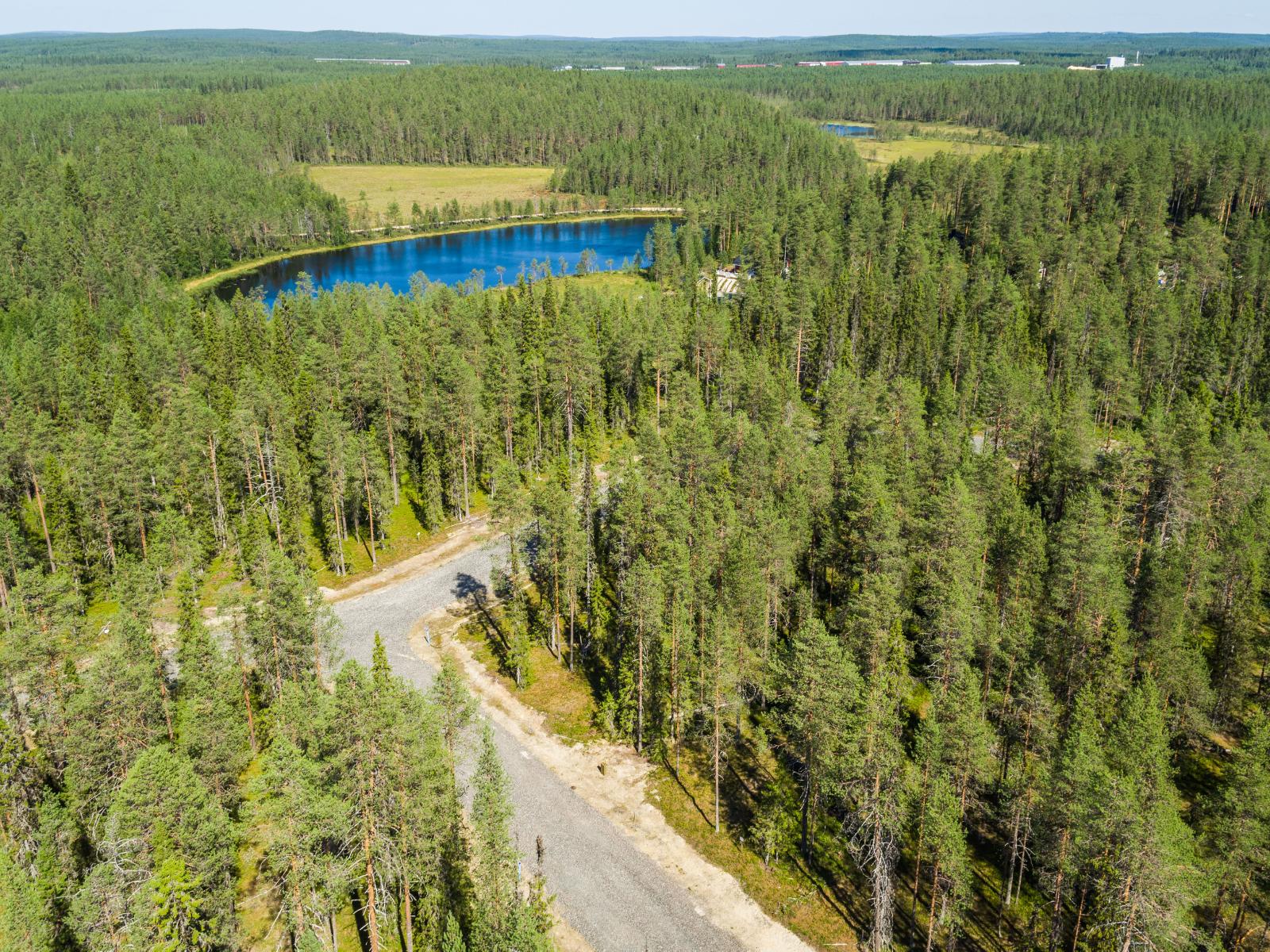 Kesäisessä ilmakuvassa tie haarautuu havumetsässä. Lähellä horisonttia on vesistöjä ja peltoja.