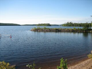 Kesäisessä järvimaisemassa aallonmurtaja rajaa veneenlaskupaikan ja vastarannalla on sekametsää.