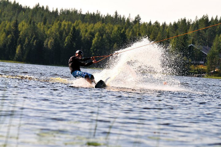 Vesi roiskuaa, kun wakeboard-harrastaja viilettää järvellä. Rantametsässä on mökki.