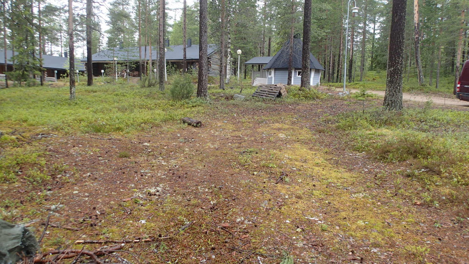 Ylävasemmalla hirsimökkejä ja kota mäntykankaalla. Etualalla on paljas tienpohja.