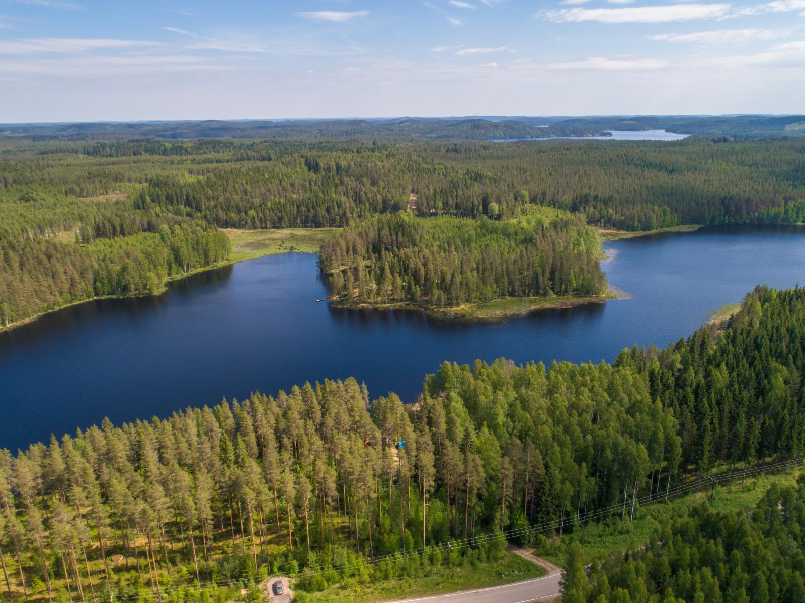 Metsä reunustaa pitkulaista järveä molemmin puolin. Etualalla on tie ja rannassa mökki. Ilmakuva.