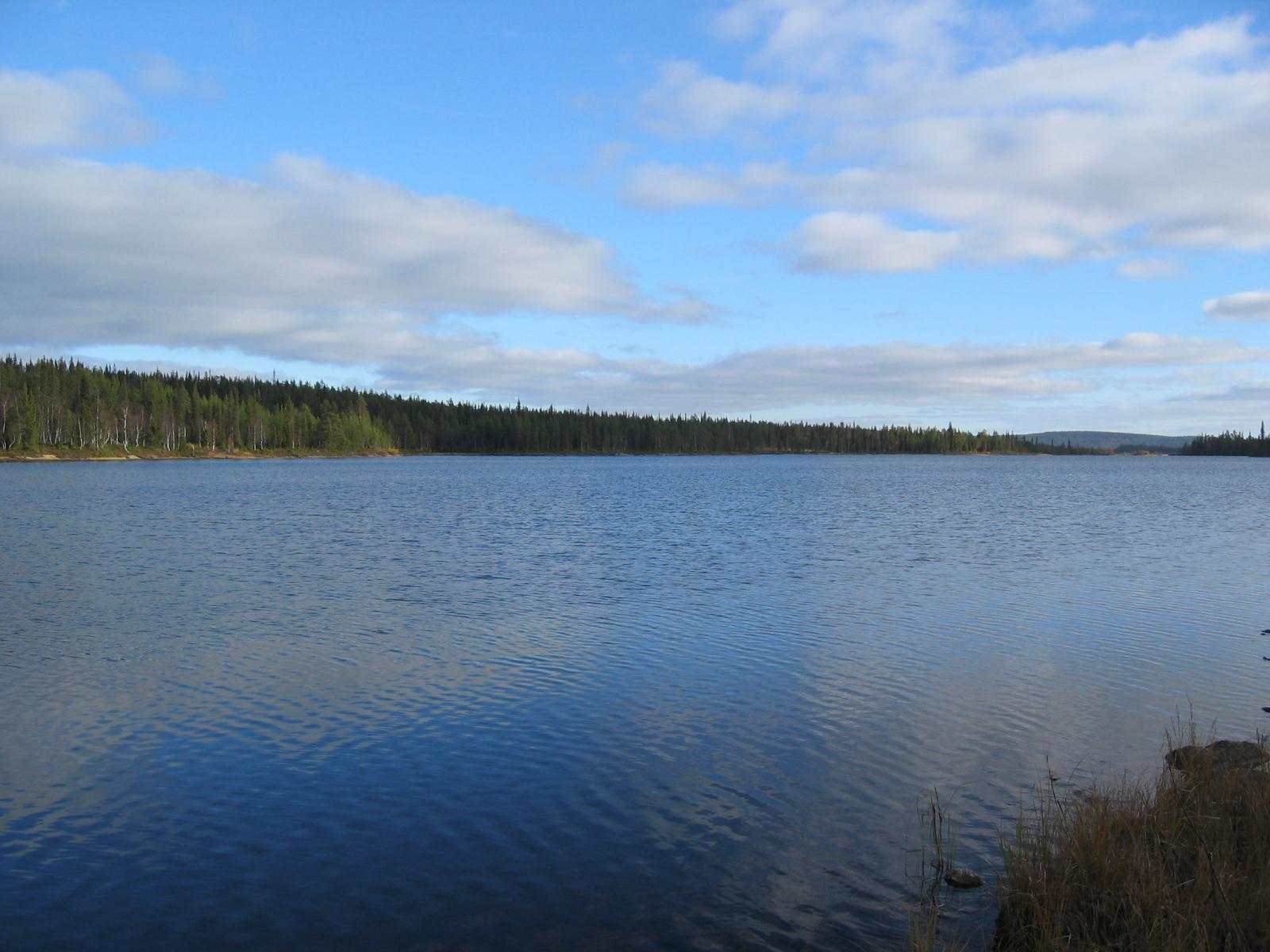 Kesäisessä järvimaisemassa puolipilvinen taivas heijastuu veteen. Vastarannalla kasvaa sekametsä.