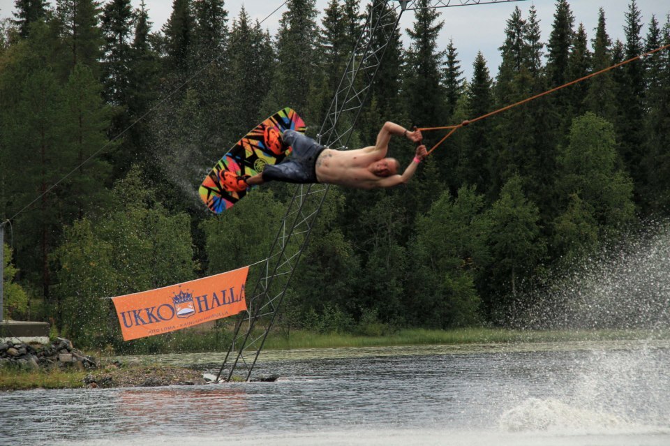 Wakeboard-harrastaja tekee ilmatemppua korkealla järven yläpuolella. Taustalla Ukko Hallan banneri.
