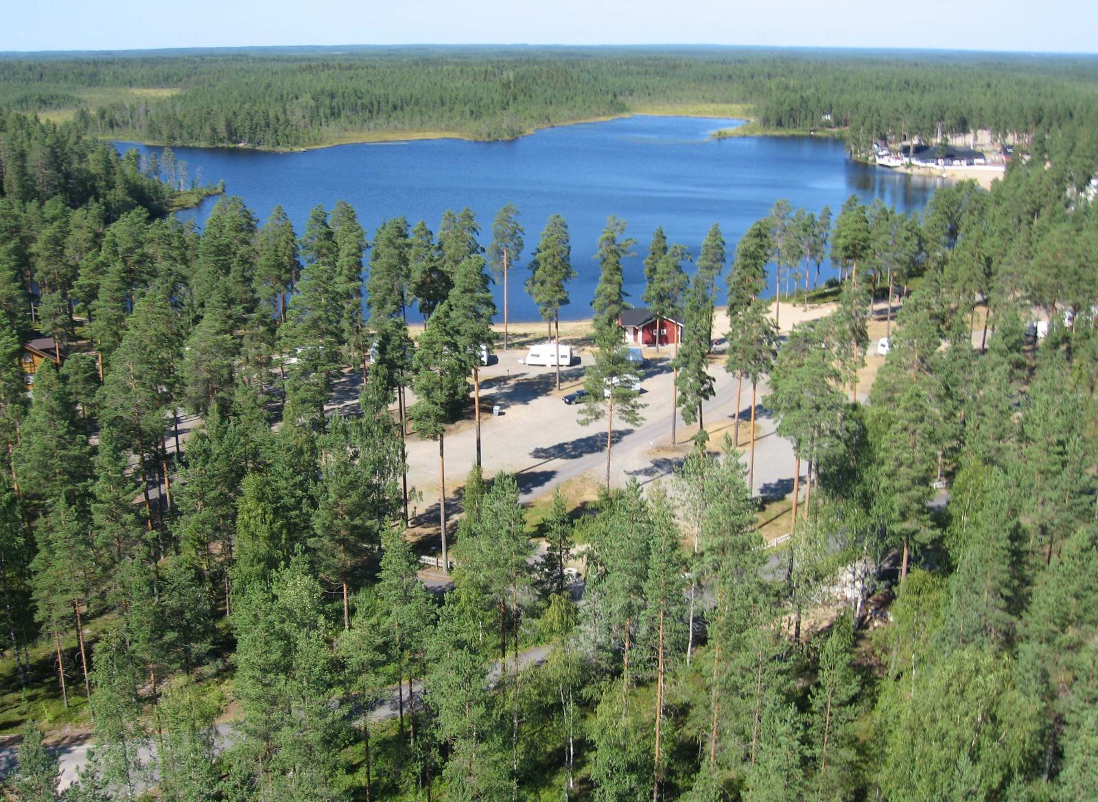 Etualan mäntymetsässä on leirintäalue järven rannalla. Vastarannalla on metsää. Ilmakuva.