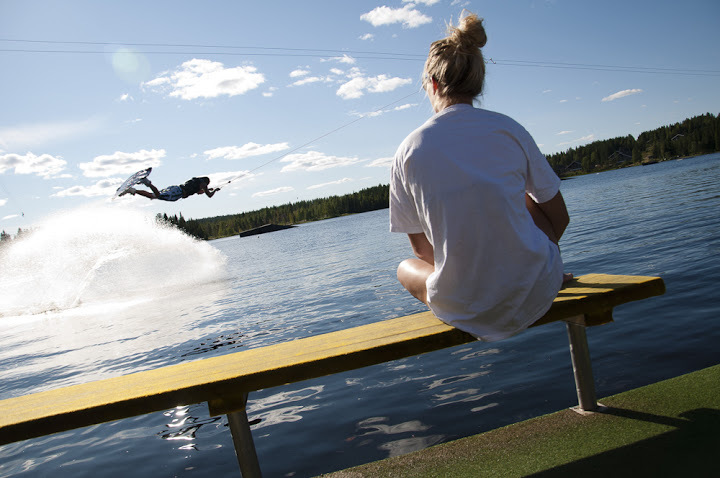 Rantapenkillä istuva lomailija katsoo wakeboard-harrastajan temppua järvellä kesäpäivänä.