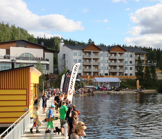 Useita ihmisiä järven yli vievällä kävelysillalla. Vastarannalla matkailukeskuksen rakennuksia.