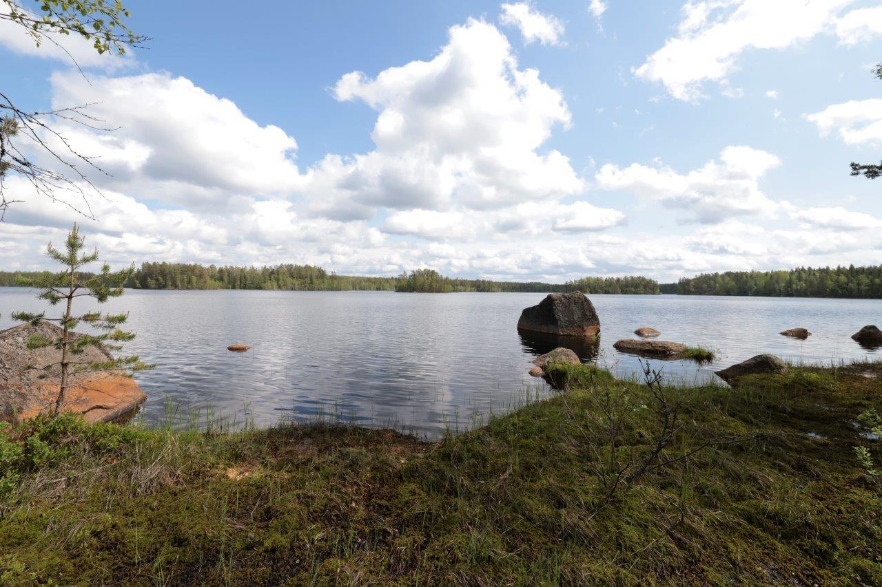 Kesäisessä järvimaisemassa puolipilvinen taivas heijastuu veteen. Rantavedessä on suuria kiviä.