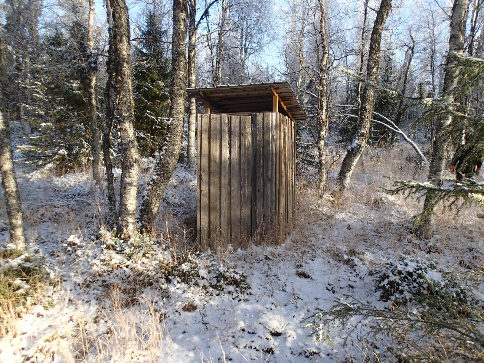 Koivut ympäröivät vanhaa huussirakennusta lumipeitteisessä metsässä.