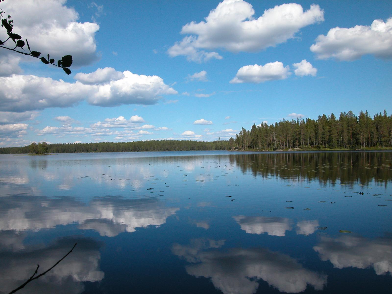 Sinisen taivaan poutapilvet heijastuvat järven tyyneen pintaan. Vastarannalla on metsää.
