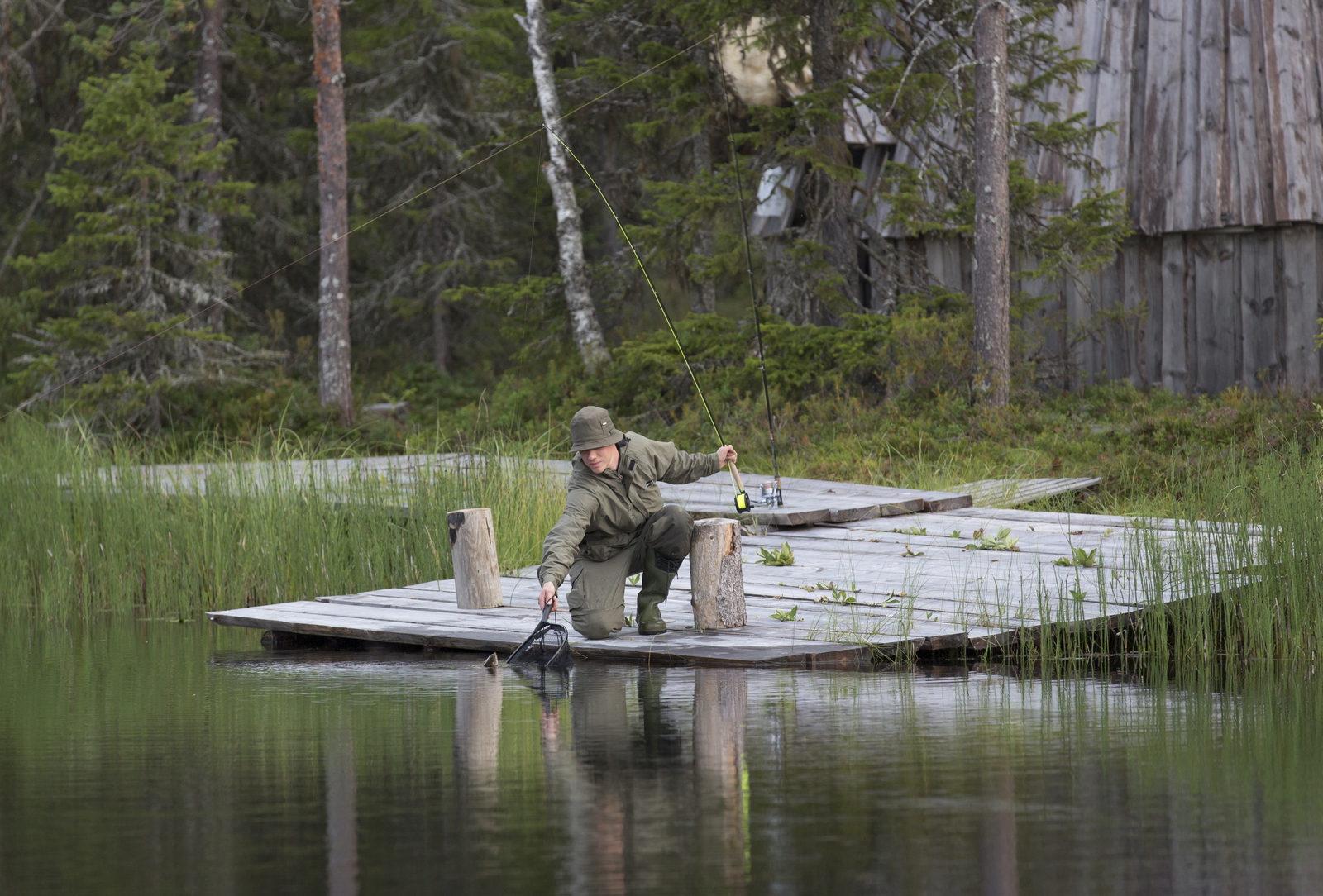 Kyykistynyt kalastaja nostaa haavilla saalistaan laiturin päädystä. Rantametsässä on rakennus.