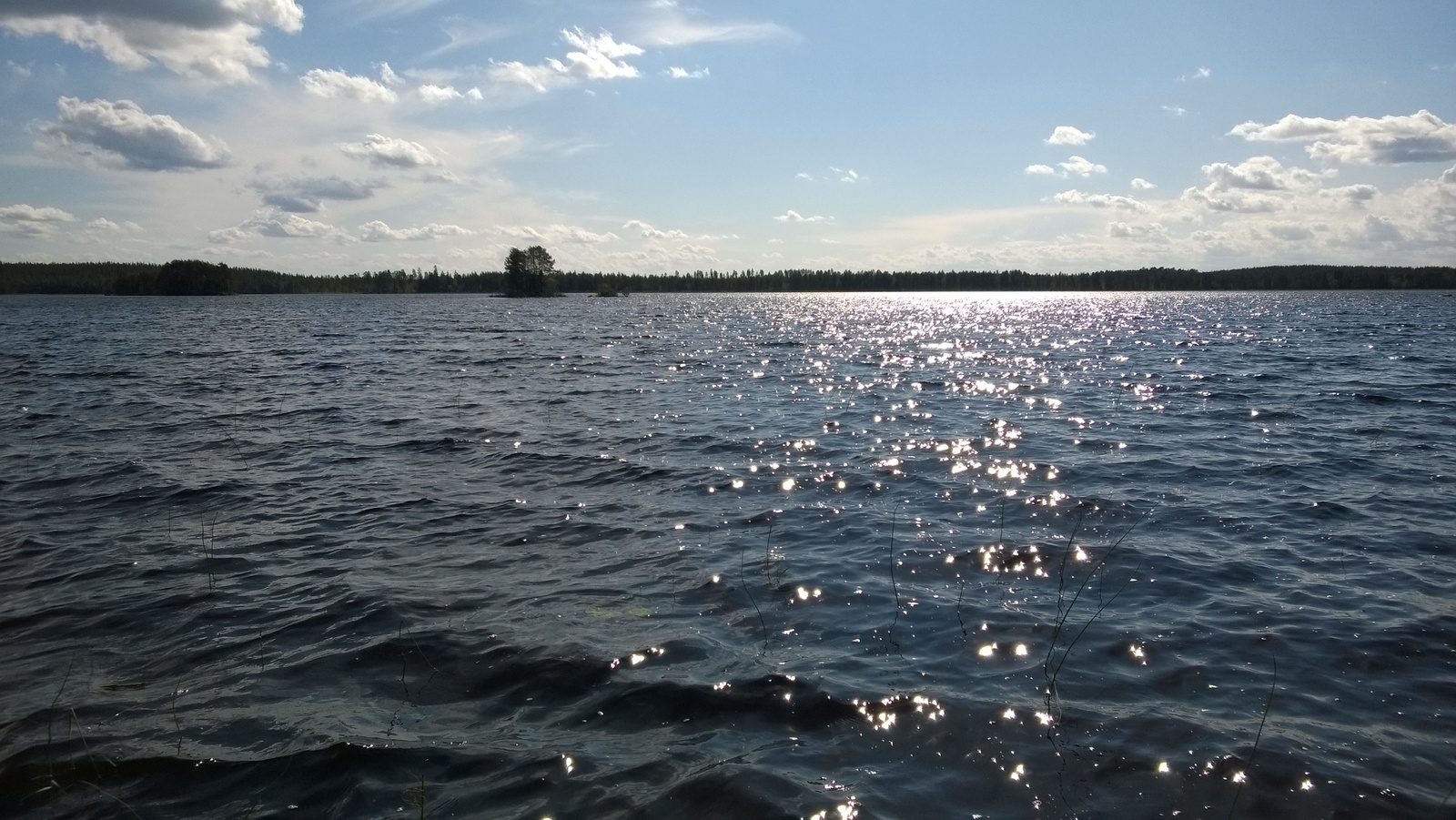 Kesäisessä järvimaisemassa laineileva veden pinta kimmeltää auringossa. Taivas on puolipilvinen.