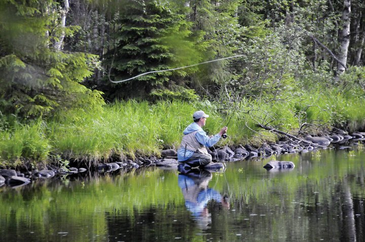 Heittokalastaja rantakivellä. Rantametsä heijastuu tyyneen vedenpintaan.