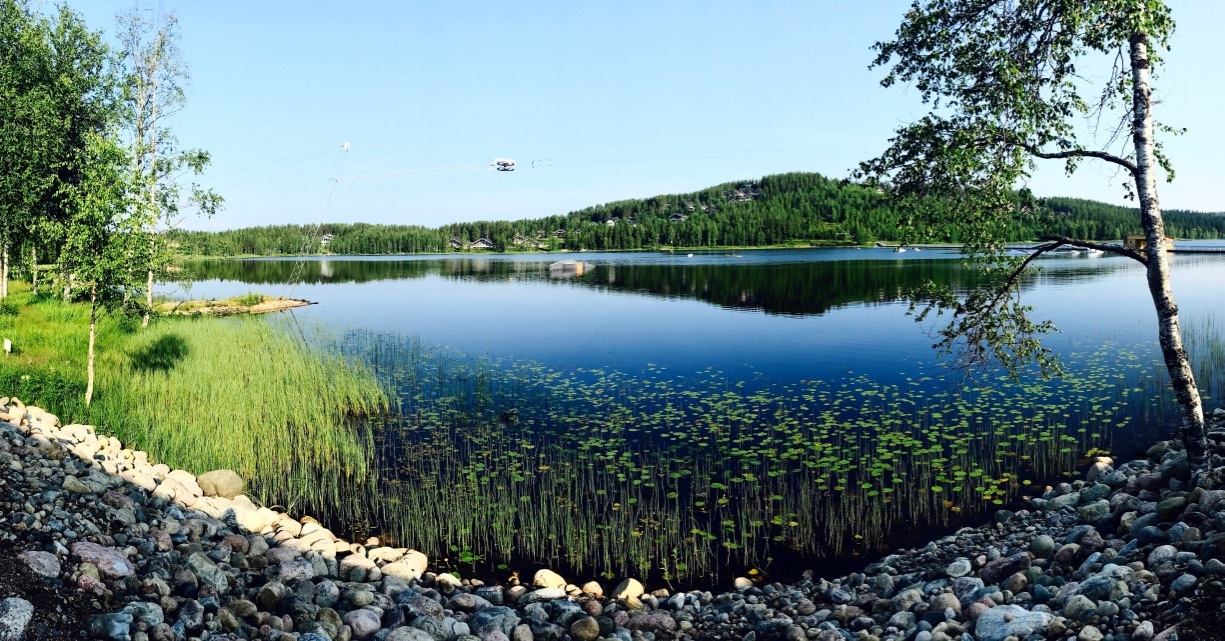 Rantakivikolta avautuvassa järvimaisemassa on vesikasvillisuutta ja vastarannalla kohoaa rinne.