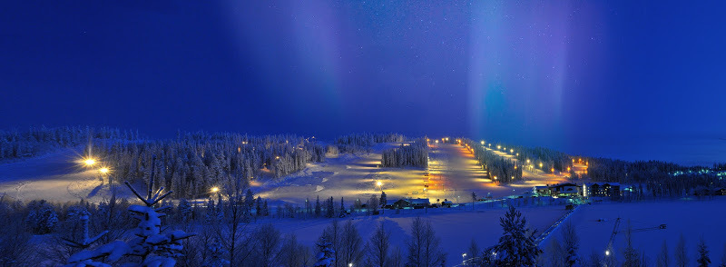 Talvimaisema valaistuihin laskettelurinteisiin, joiden yllä revontulet loimuavat.