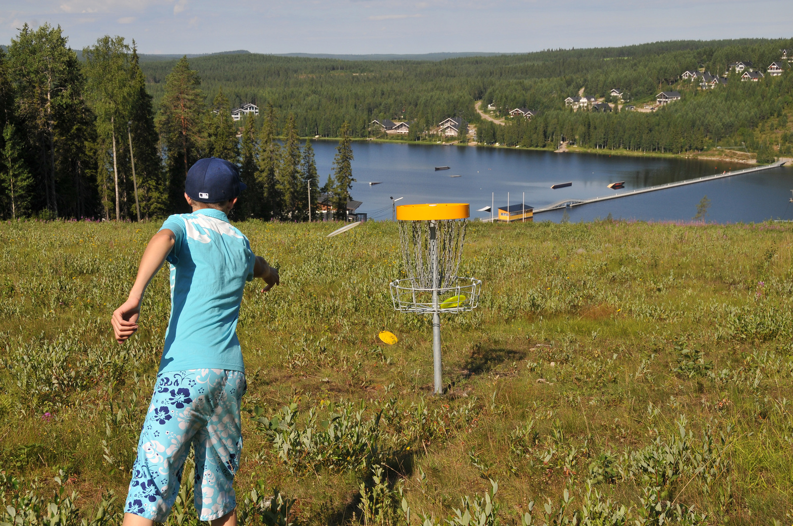 Lapsi pelaa frisbeegolfia korkealla rinteessä. Alhalla olevan järven rannalla on mökkialue.
