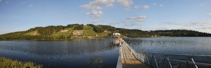 Kesäinen järvimaisema. Kävelysilta vie järven yli kohti vastarannalla kohoavia laskettelurinteitä.