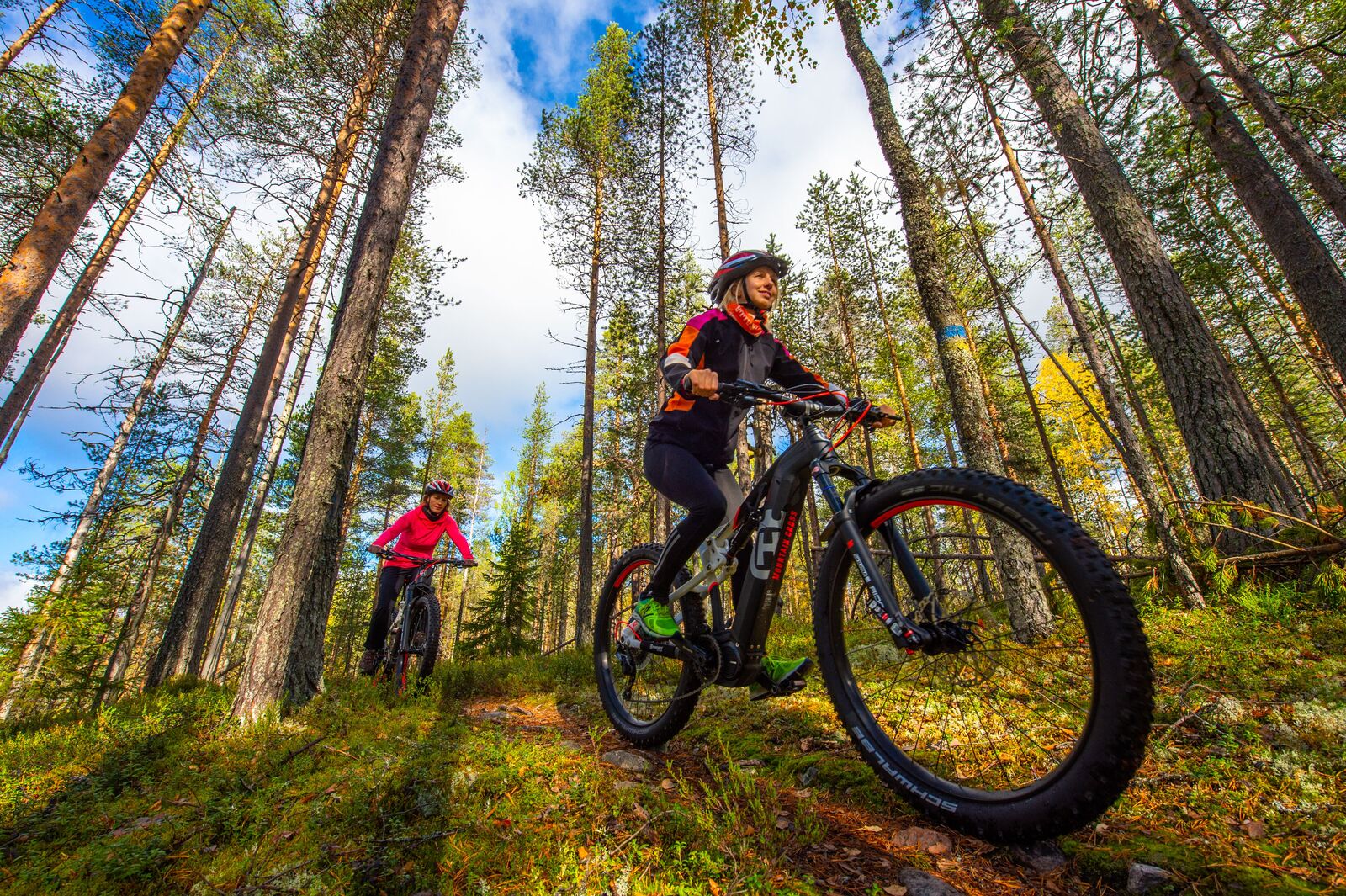 Hymyilevät maastopyöräilijät polkevat merkityllä reitillä valoisassa metsässä.