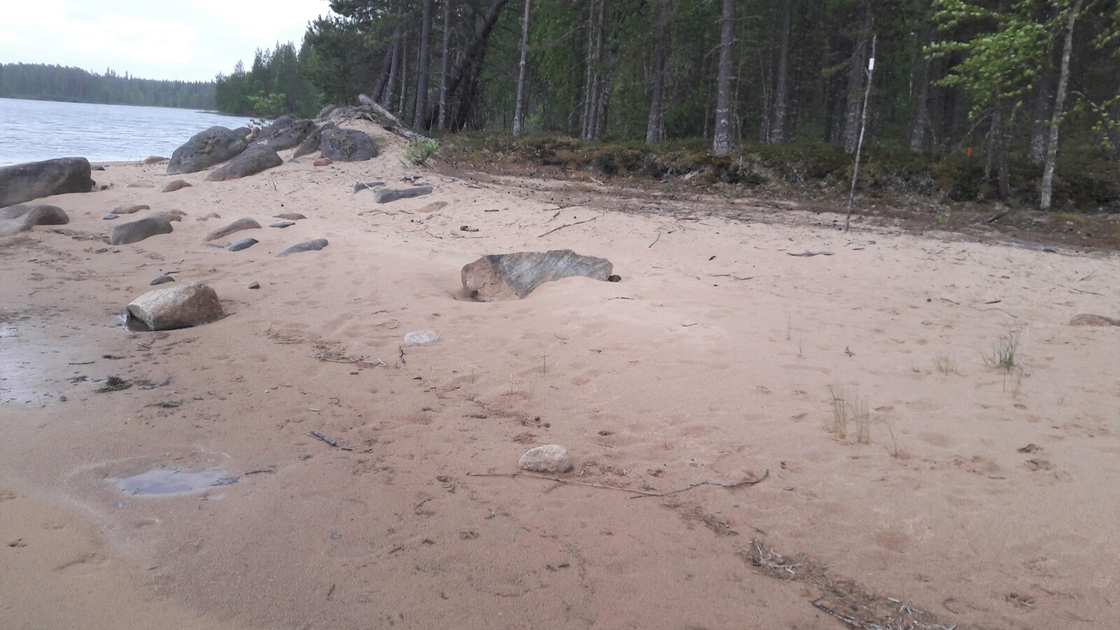 Etualan hiekkarannalla on kiviä. Taustalla on järvi, jota reunustaa sekametsä.