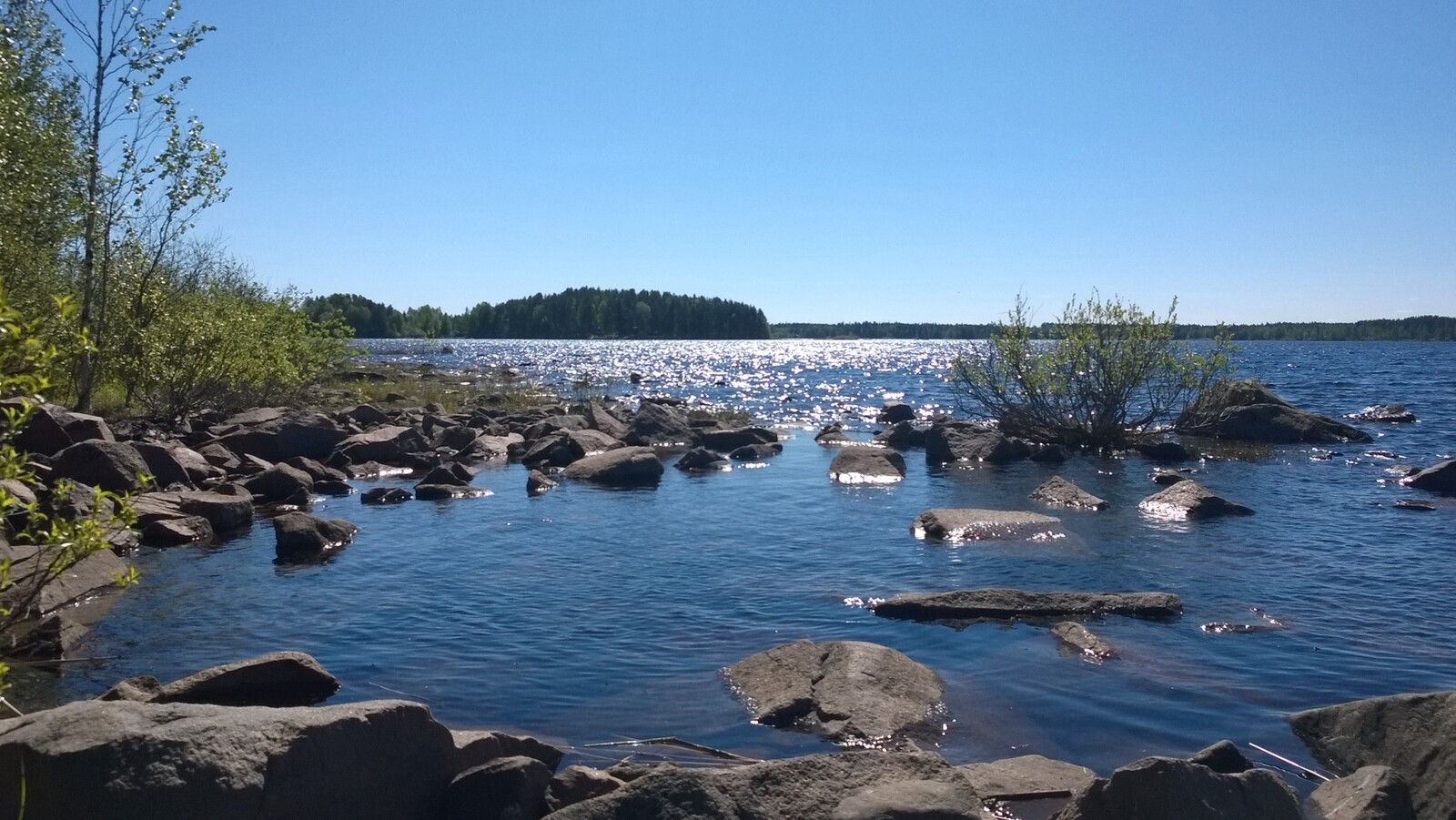 Rantaveden kivikosta aukeaa kesäinen järvimaisema. Veden pinta kimmeltää auringonvalossa.