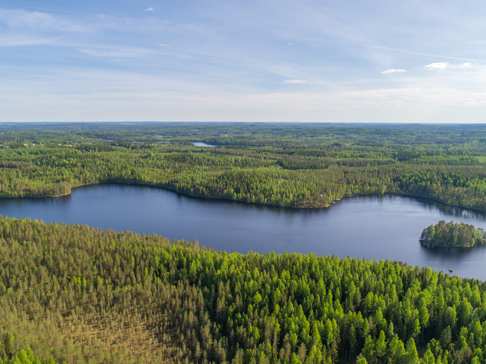 Kesämaisemassa laajat metsäalueet reunustavat pitkulaista järveä. Ilmakuva.