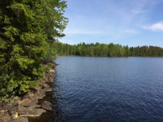 Sekametsä reunustaa kesäistä järvimaisemaa oikealle kaartuvassa kivisessä rantaviivassa.