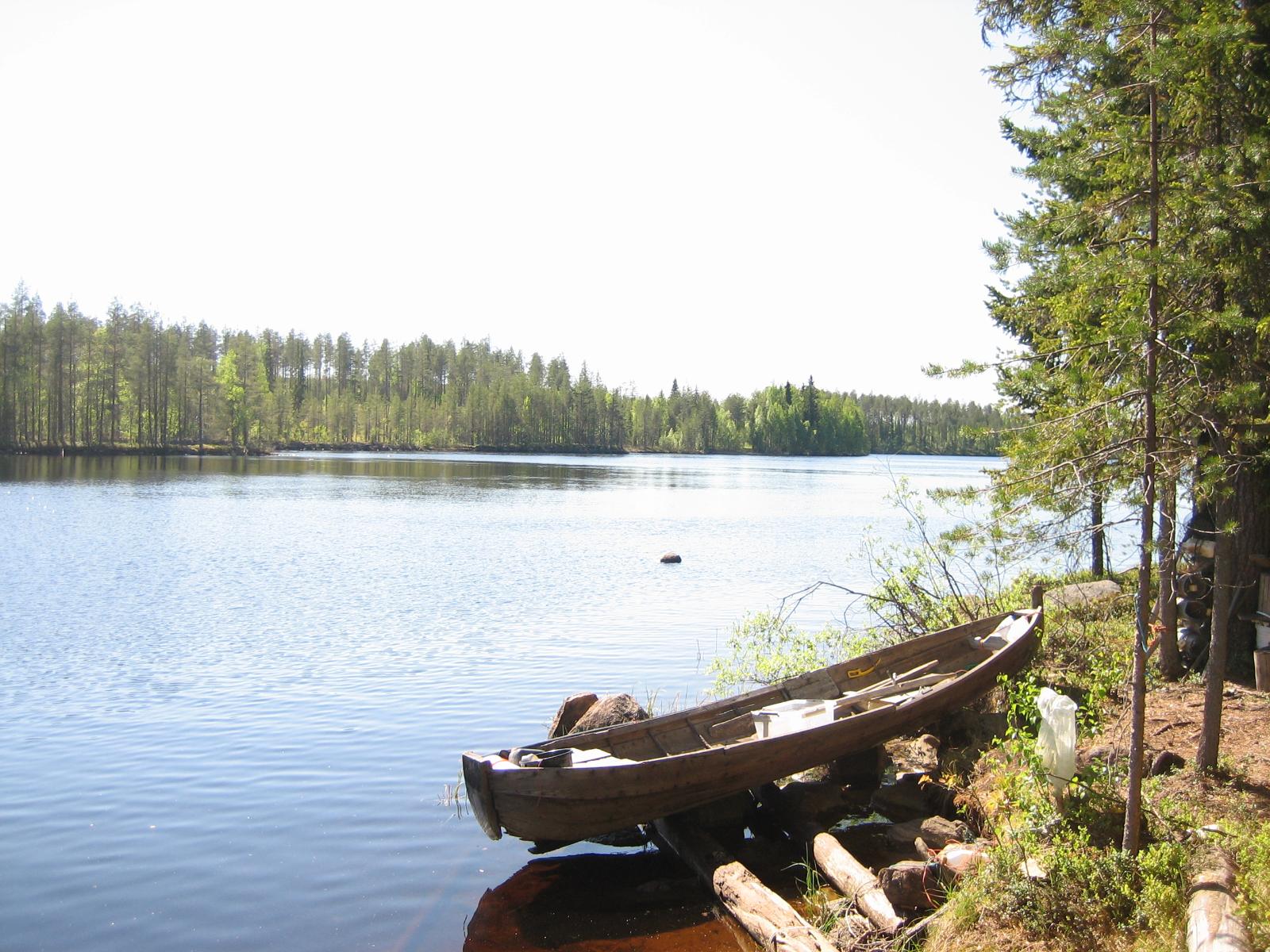 Kesäisessä järvimaisemassa puinen vene on vedetty maihin oikean laidan rantaan. Rannalla on mäntyjä.