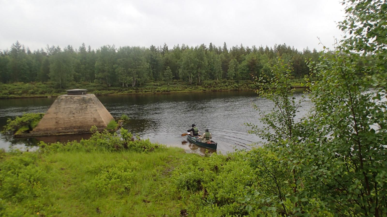 Rannan tuntumassa on kaksi melojaa kanootissa. Vastarannalla jokea reunustaa metsä.