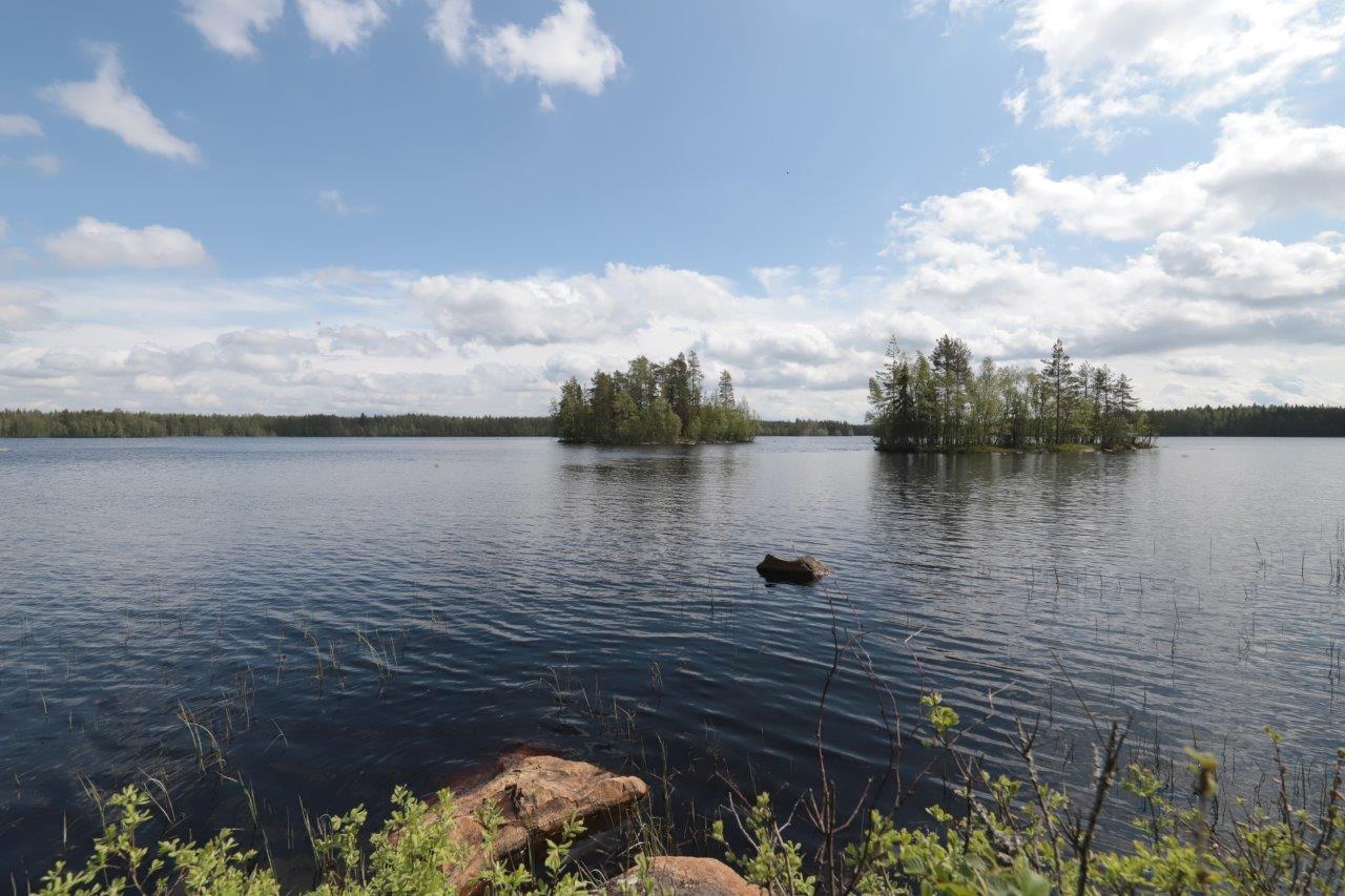 Kesäisessä järvimaisemassa puolipilvinen taivas heijastuu veteen. Järvessä on kaksi saarta ja kivi.