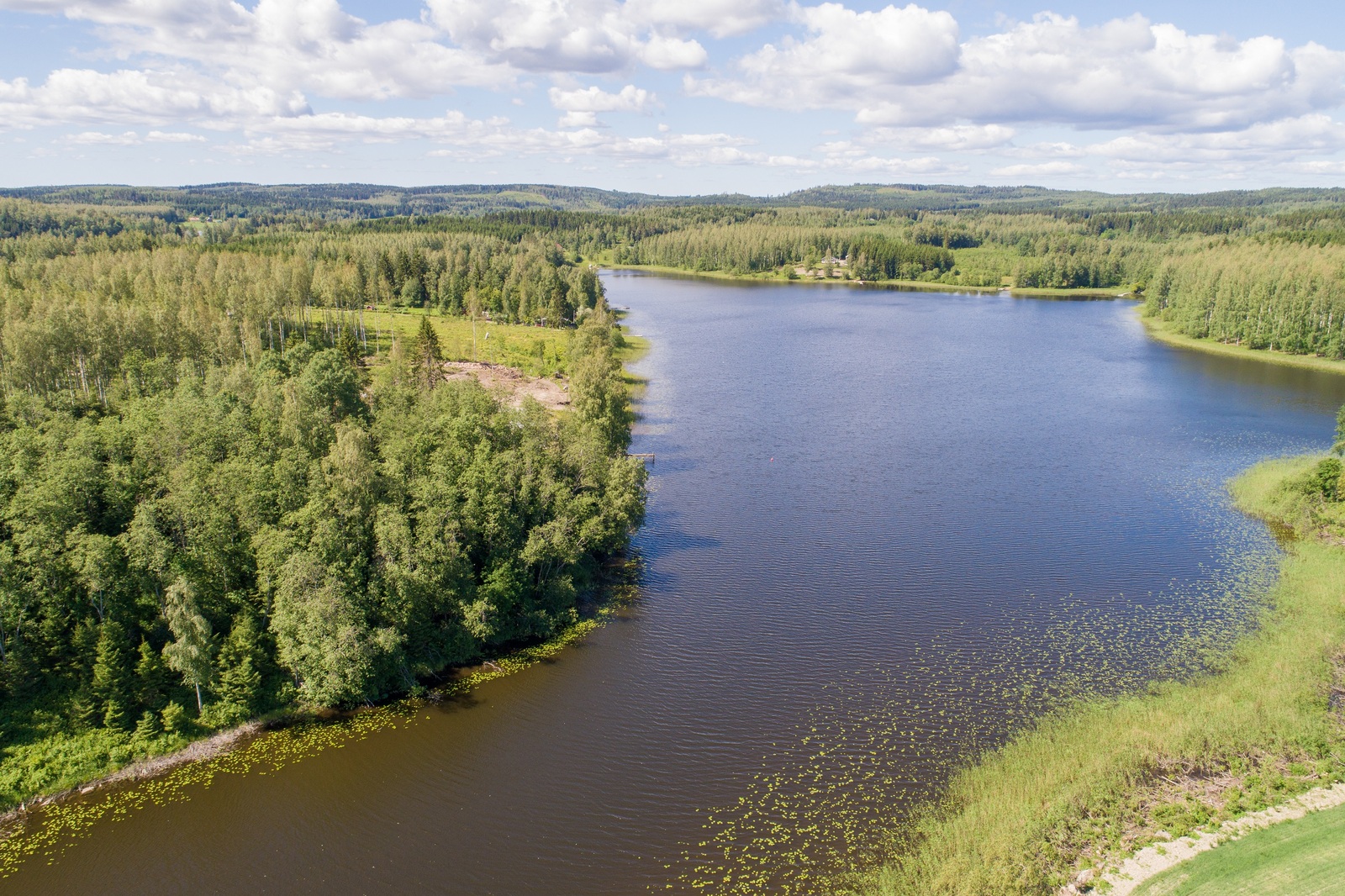 Kesämaisemassa metsät reunustavat pitkulaista järveä. Rannalla on rakennuksia. Ilmakuva.