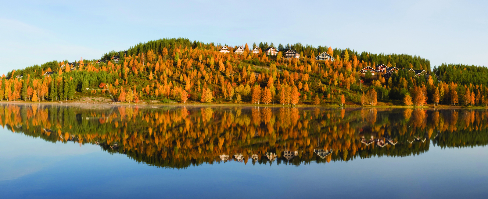 Tyyneen järven pintaan heijastuu ruskan väreissä hehkuva rinnemaisema. Rinteessä on mökkejä.